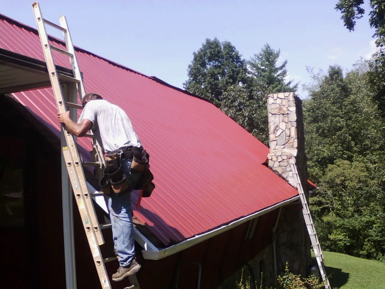 Roofer on a ladder beside a red metal roof and stone chimney — Soffit & Fascia Repair by Roofing Brothers in Centerport